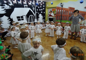 Na tle zimowej dekoracji dzieci przebrane za gwiazdeczki wykonują taniec trzymajc ręce na biodrach.